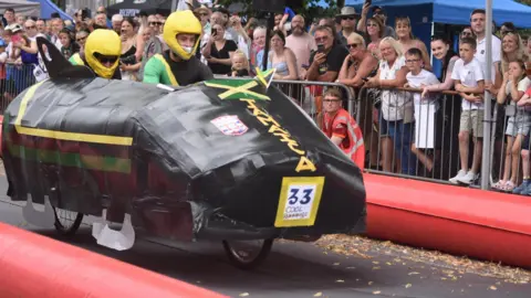Rachel Wield A large black boblsed with yellow and green stripes down the side. On the front, vertical text reads JAMAICA.  Theres two riders inside wearing yellow helmets.