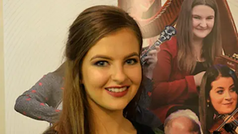 Ashling Murphy standing in front of and Irish music sign. She has long brown hair and blue eyes.