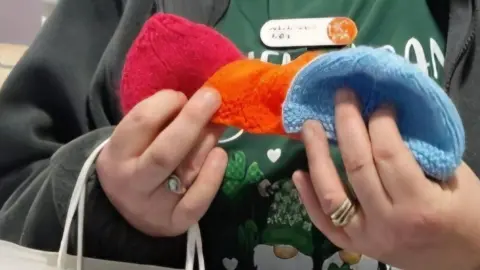 HC One A close-up of a woman's hands holding a set of knitted hats. There is a red one, and orange one and a blue one. The woman is wearing a green t-shirt and dark grey jumper.