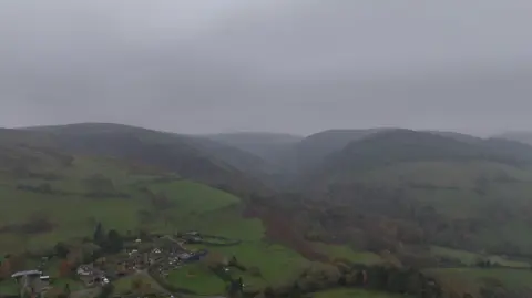 A foggy hillside scene, viewed from the air, with large hills in the distance and some properties in the foreground