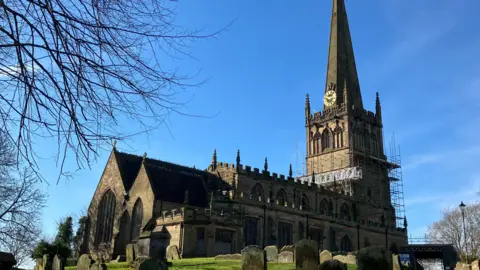 A 12th-century church with graves in the foreground on green grass, set against a blue sky. The spire is covered in scaffolding.