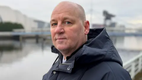 A bald man stares directly at the camera with a serious expression on his face. He is wearing a blue hooded anorak. A river and buildings can be seen blurred in the background.