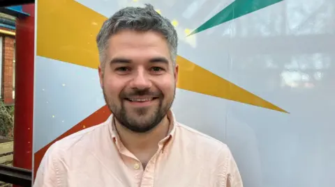 George King/BBC Jake Smith is standing in front of the Eastern Angles logo, which consists of coloured triangles on a white background. Jake is wearing a peach shirt, has a beard and is smiling at the camera. 