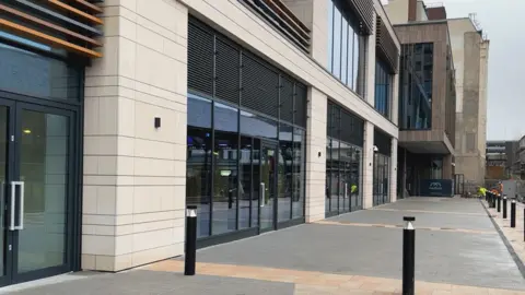 A close up of the ground floor entrance showing huge dark glass doors with black casings, surrounded by sandstone. The pavement is smooth grey flags, broken up with paler flags in stripes. There are black safety barriers around it