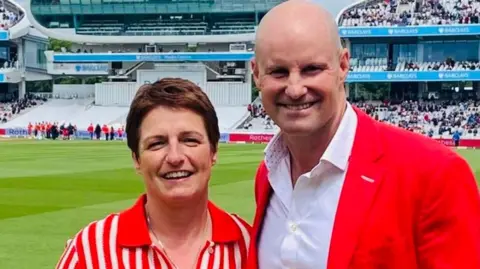 A woman and a man (former England cricket captain, Sir Andrew Strauss) are standing on Lords cricket pitch. The woman is dressed in a red and white blouse and has short hair. The man is dressed in a white shirt and red blazer and has no hair. There are spectators in the stands behind them.