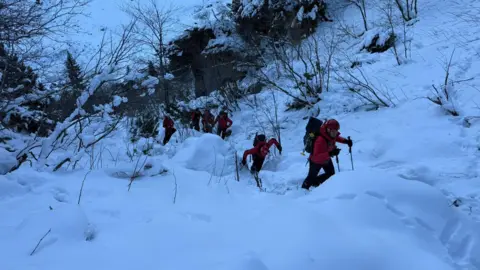 Salvamont Brașov A group of rescue personnel wearing red jackets, black backpacks and holding black walking poles are walking through deep snow. There are snowdrifts and piles around them, with bare trees and bushes all around