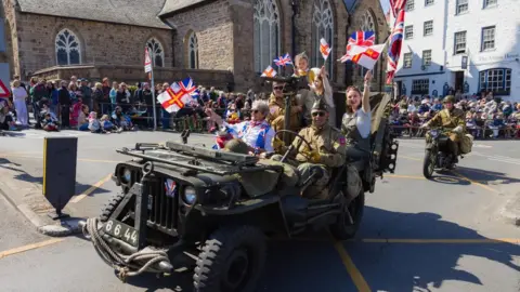 People sat in a green vehicle. They are waving Guernsey flags and there is a crowd of people around them. There is a motorbike following the vehicle.
