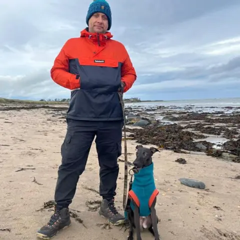 Corey Nobbs A man stands next to his dog, a small black whippet on a beach next to the sea. The dog is wearing a blue dog coat, with orange on it. The man is wearing a red and blue rain jacket, blue trousers and walking boots. Behind them, seaweed and rock pools can be seen under a grey, cloudy sky.