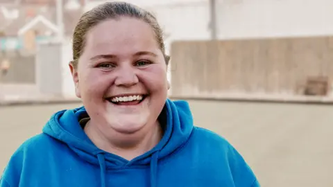 Jo-Anne looks directly into the camera, wearing a bright blue hooded top and her hair up in a bun.