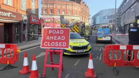 Stafford Street in Hanley, with a sign saying road ahead closed. Behind red barriers a police car and fire engine is visible. A large red brick property can be seen in the distance, with other shops also visible. 