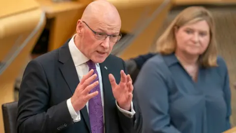 John Swinney, who has a bald head and glasses, speaks in the Scottish Parliament chamber. He is wearing a dark suit, white shirt and purple tie. 