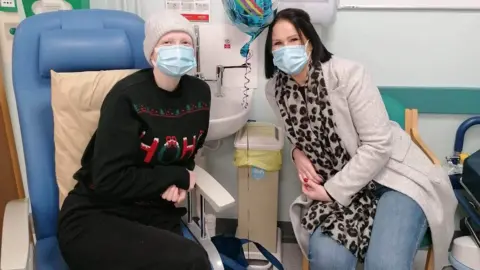 University of Salford Lucy Wiswould-Green sits in a chair during treatment for cancer, with her mum on a chair next to her. Both are wearing masks 