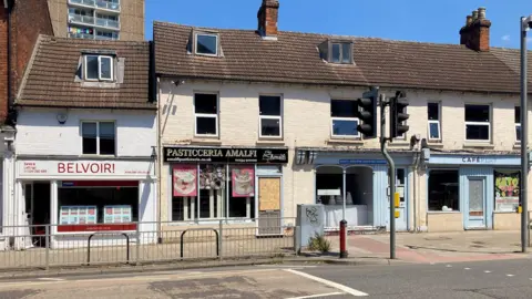 A terrace of shops pictured on a sunny day. They include an estate agency, restaurant and cafe. They all front onto a road.
