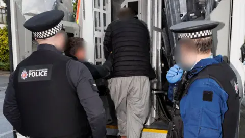 A man with his back to the camera is helped into the back of a police van by two uniformed officers. 