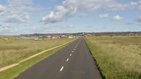 Google A  view along La Route de la Pulente. it is a long straight road with grass either side of it. There are cars on the road in the distance. 