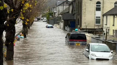 Cars stranded in flooded street as man uses a bucket to tip water over wall