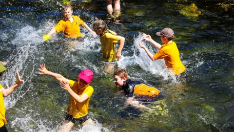 A handful of children in a shallow stream splash each other. They are wearing yellow school polos and baseball caps.