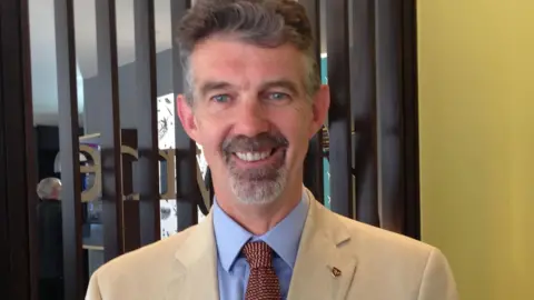 Skelly, sonriente, con chaqueta beige, camisa azul y corbata roja en un evento de negocios.