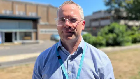 George Carden / BBC a man wearing a pale blue shirt and a lanyard around his neck faces the camera as he stands in the sunshine. He has short white hair, white stubble and is wearing colourless framed glasses.
