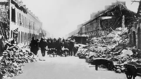 Getty Images Rubble is on the sides of a street. Houses are damaged and brick and timber are exposed. People are trying to clear some of the debris. 