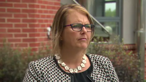 BBC A woman, Tina Graham, with medium length brown hair, in glasses wearing a black and white jacket and a flowery silver necklace