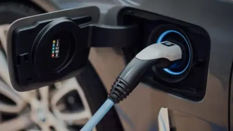 A car is being charged at an electric charging point. The plug can be seen in the socket built in to the side of the vehicle