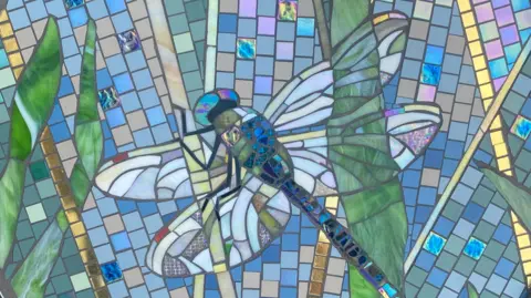 A close-up of the mosaic shows a southern migrant hawker dragonfly. The tribute is made up of gold and multi-coloured pieces of glass.