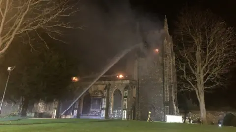 BCH Road Policing Firefighters spray a hose at at St John the Baptist Church, Royston, while flames flicker on the roof at night.
