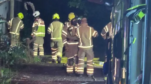 Harpenden Fire Station Five firefighters, one wearing breathing apparatus, stood by a fire engine. They are wearing being safety equipment with fluorescent strips 