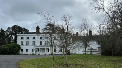 Picture shows the Georgian house with its white facade and uniform windows around a central door and canopy with columns.
In front is a sweeping driveway and grass lawns with young trees