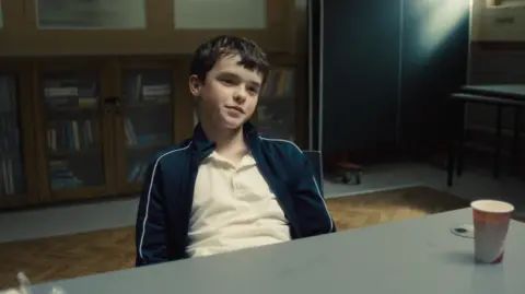 Netflix A young boy sitting on a chair in front of a grey table with a takeaway cup on the right. The boy is dressed in a navy tracksuit jacket and white polo shirt. 