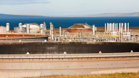 A large oil terminal site with various buildings and storage tankers sitting next to a wide expanse of sea with land in the distance