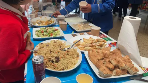 A display of food laid out on a table with people around it. There is a rice, bread, salad and pastries