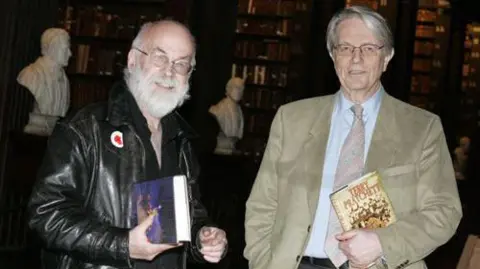 Trinity College Dublin Sir Terry is dressed in a black leath jacket next to Colin Smythe who is wearing a tan coloured blazer and blue shirt. They are both holding hardback copies of his book and are stood together in a library