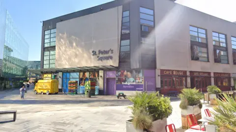 An open, concrete floor space with restaurant table and chairs to the bottom right. A grey building with black-framed buildings is in the background.
