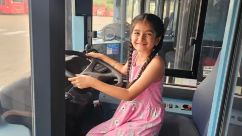 A young girl sits in the drivers seat of a bus in a pink dress 