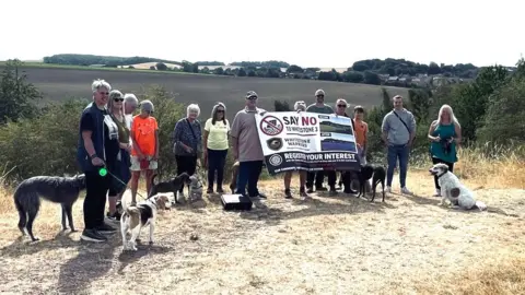 Paul Rowland A group of people are stood by open fields and countryside, they have a large banner objecting to plans for a solar farm