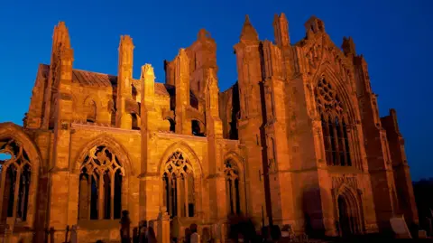 Melrose Abbey pictured at night light up by floodlighting, it is an imposing sandstone building with various towers jutting up into the dark sky