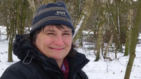 Dawn Balmer A woman is wearing a blue coat and a blue woolen hat. She is standing in front of a number of small trees and there is snow on the ground. 