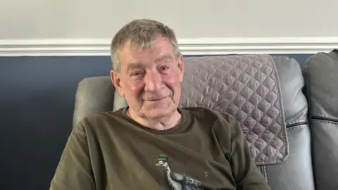 Bob is sitting on a grey sofa and smiling. He is wearing a green top and has sandy grey speckled hair. 