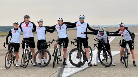 A team of seven male cyclists in black and white clothing standing astride their bikes on a runway of an airport. 