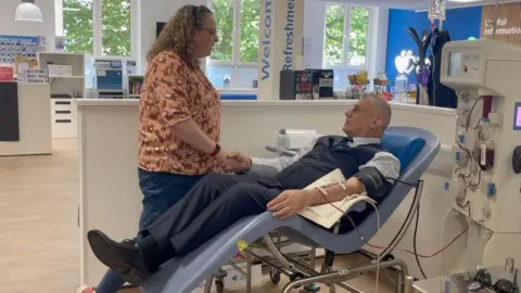 BBC A man and woman shaking hands. The man is lying down a blood donation bed. He is donating plasma so is attached to a machine. The woman is standing next to the bed. She is wearing an orange top and has curly hair.