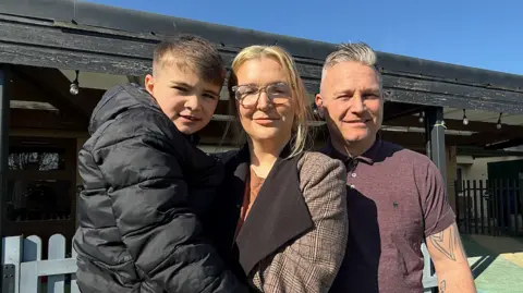 Tori Hetherington who has blonde hair, wearing clear glasses and a chequered coat, is holding her son, Nate, who has black hair and is wearing a black puffer jacket. Tori is standing next to her partner, Lee, who has grey hair and is wearing a purple short-sleeved shirt.