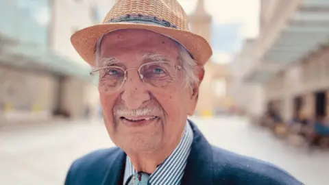 Yavar Abbas with white hair and moustache, looks at the camera wearing glasses and a straw hat.