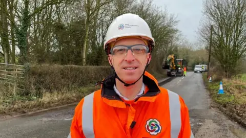 Ollie Conopo/BBC Peter Ingram wearing a white hat and glasses looking at the camera with orange hi-vis. A road lined with trees is behind him, and there is a yellow digger in the background