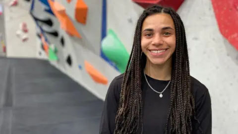 Molly Thompson-Smith pictured at her climbing gym in Sheffield. Molly is a 26-year-old woman with long braided hair worn loose. She wears a long sleeved black top and has light brown eyes. She is pictured smiling at the camera, colourful boulders and grey crash mats behind her.. 