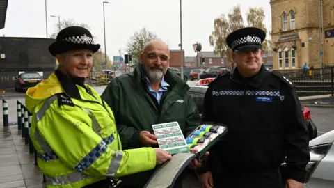 Two smiling police officers standing outside either side of Alex Chivers, a bald man with light grey facial hair, while he holds an open bag containing the RAPAID bandage kit.