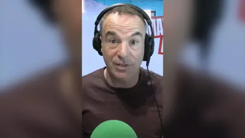 Martin Lewis wears headphones and a brown t-shirt. He sits in-front of a green microphone and looks directly at the camera.