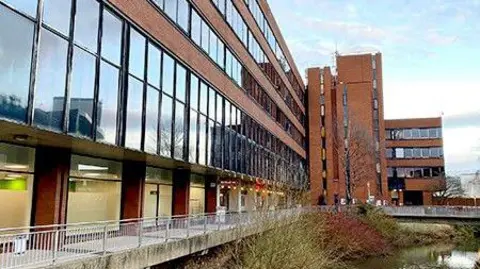 Stafford Borough Council A four-storey building with large windows overlooks a river. There is a footway alongside the building and a bridge. A number of trees and other vegetation can be seen growing along the river bank.