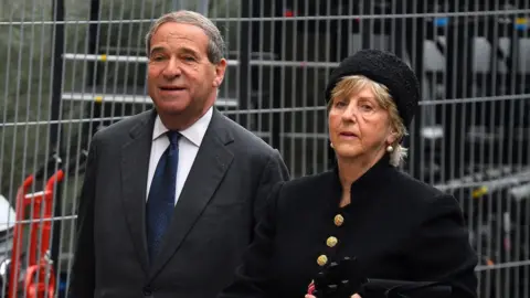PA Media Lord Leon Brittan, pictured in a suit and tie, and Lady Diana Brittan, in a matching black boucle suit jacket and hat with large gold buttons. Both are looking at the camera and are pictured while walking next to a metal fence.